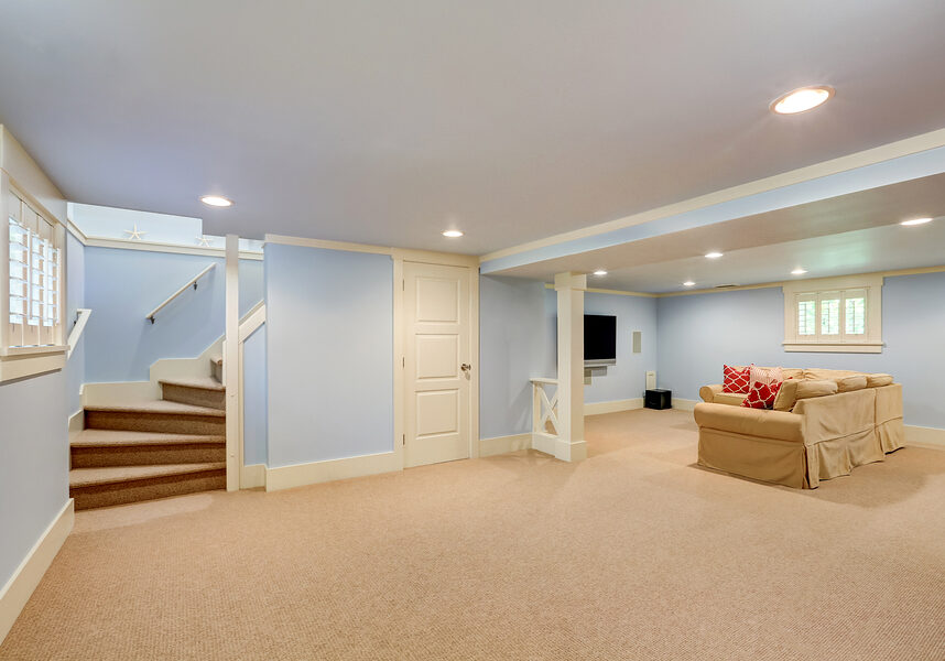 Spacious basement room interior in pastel blue tones. Beige carpet floor and large corner sofa with TV. Northwest USA